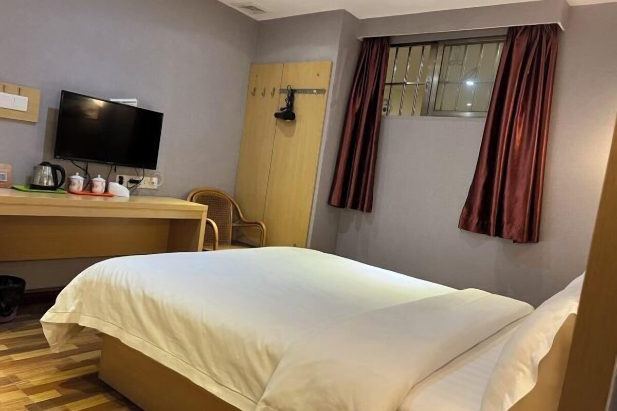 Une chambre d’hôtel avec un lit, un bureau avec une télévision, une fenêtre avec des rideaux et un plancher en bois.