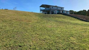 A modern two-story house with a flat roof and large windows, situated on a grassy hill with a clear sky.