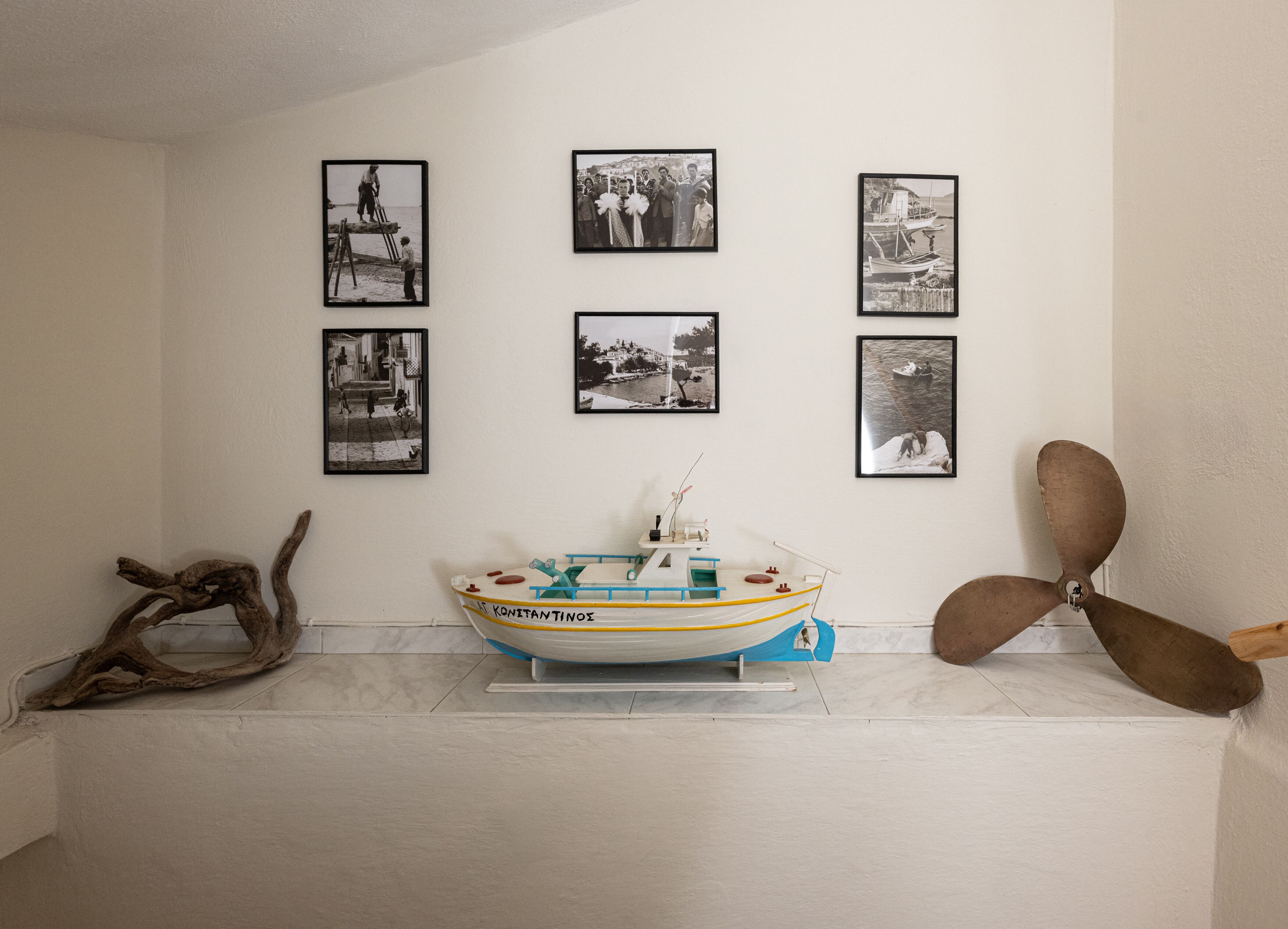 A room with a model boat on a stand, a wooden sculpture, and four black-and-white photographs on the wall.