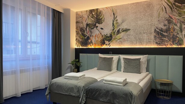 A hotel room with a bed, a plant, a wall art piece, and a window with curtains.
