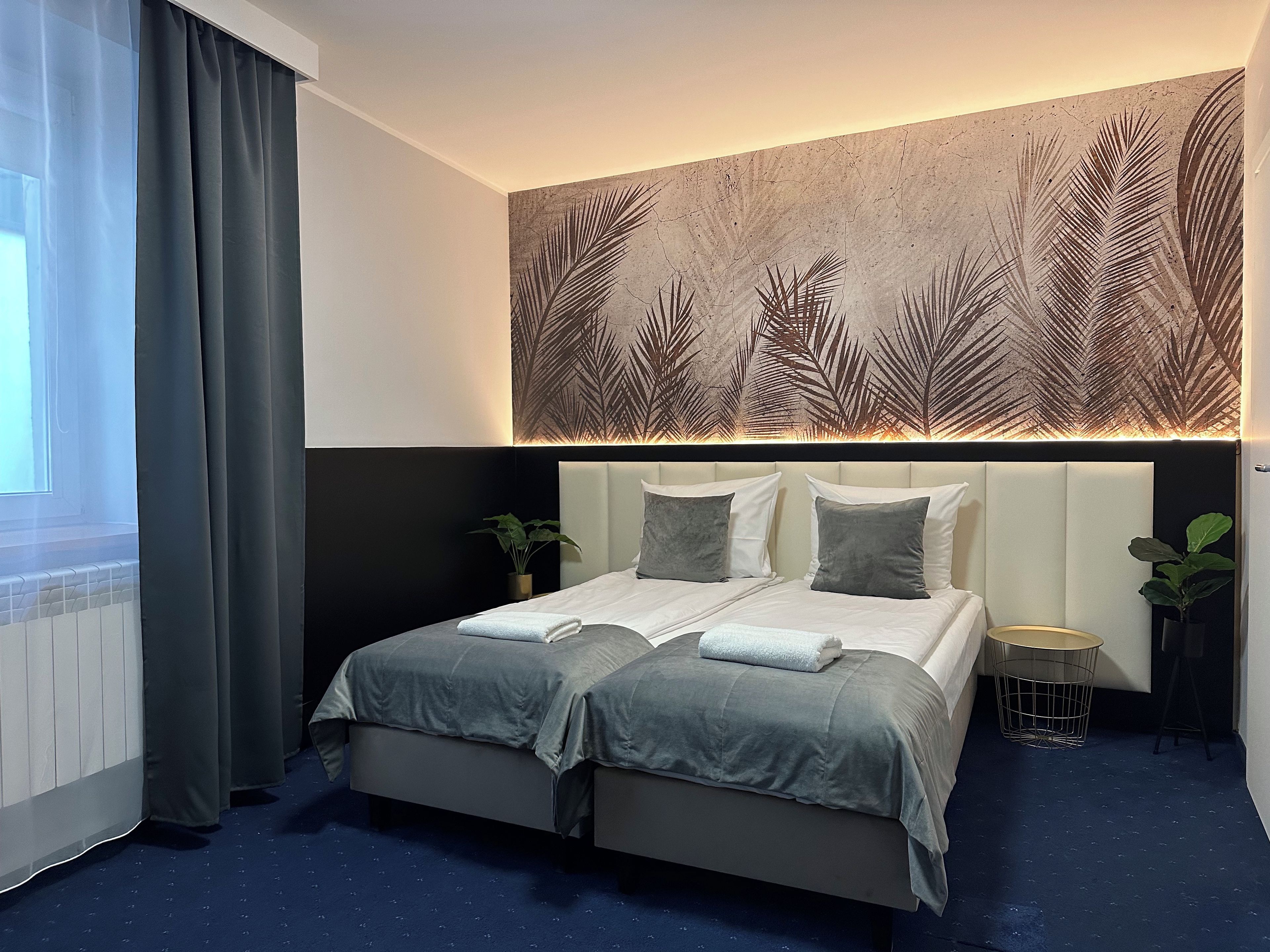 Una habitación de hotel moderna con una cama grande, alfombra azul oscuro y un mural de hojas de palmera.