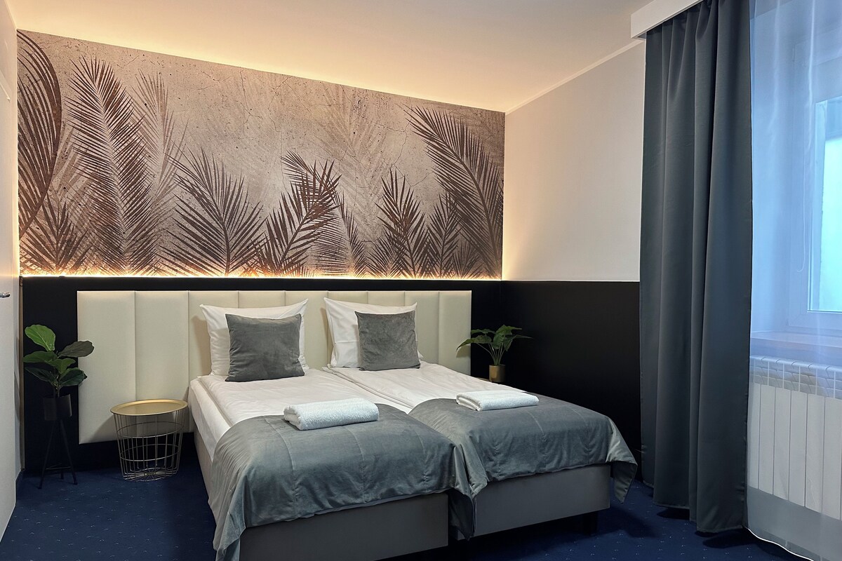 A modern hotel room with a large bed, two bedside tables, a wall-mounted light panel, and a view of the outdoors through a window with curtains.