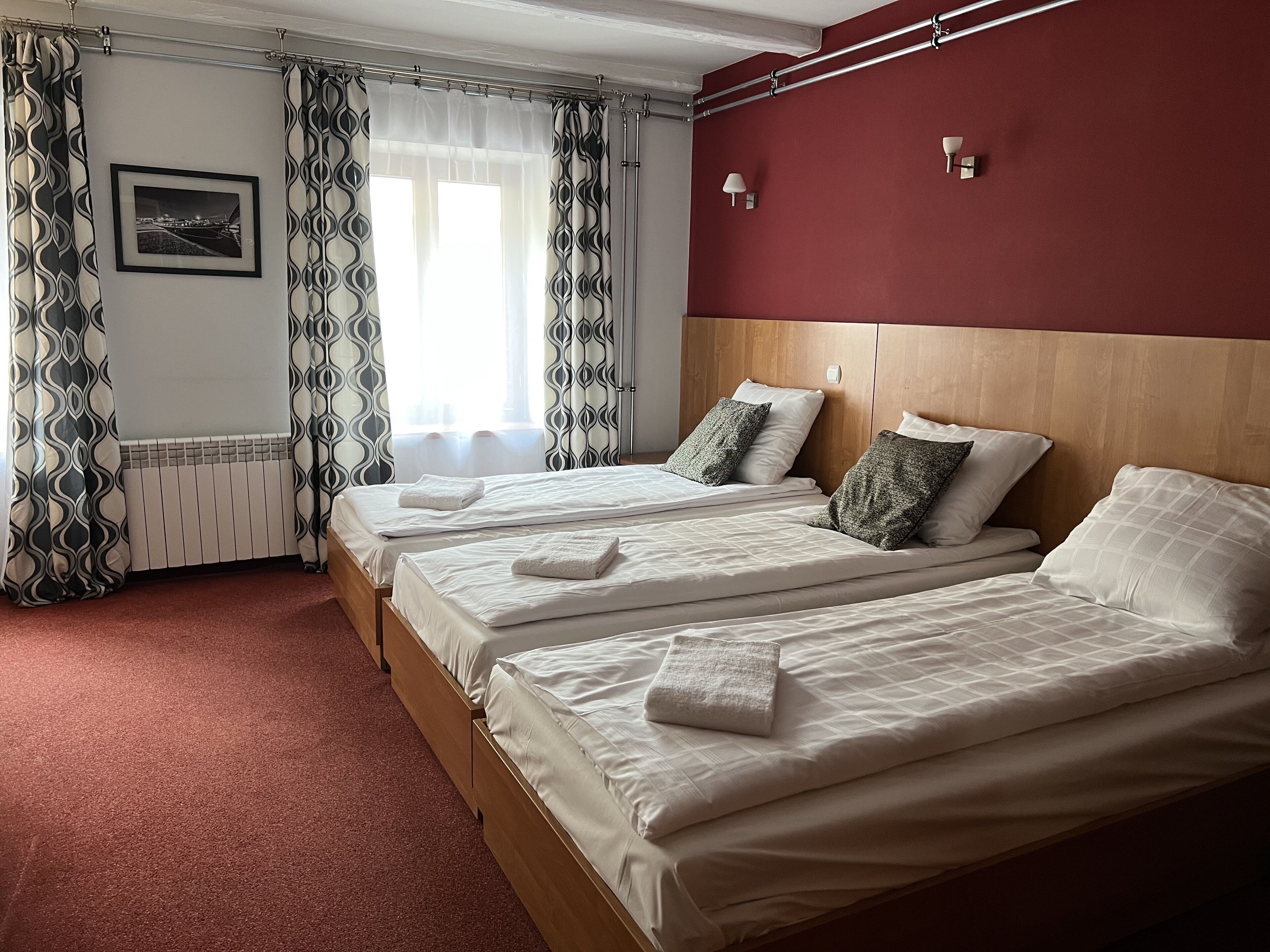 A hotel room with two beds, a red accent wall, a framed picture, and a window with curtains.