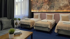 A hotel room with three beds, a large window with sheer curtains, a wall mural, and a small table with a plant and books.