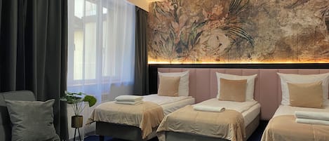 A hotel room with three beds, a large window with sheer curtains, a wall mural, and a small table with a plant and books.