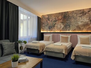 Une chambre d’hôtel avec trois lits, une grande fenêtre avec des rideaux légers, une fresque murale et une petite table sur laquelle se trouvent une plante et des livres.