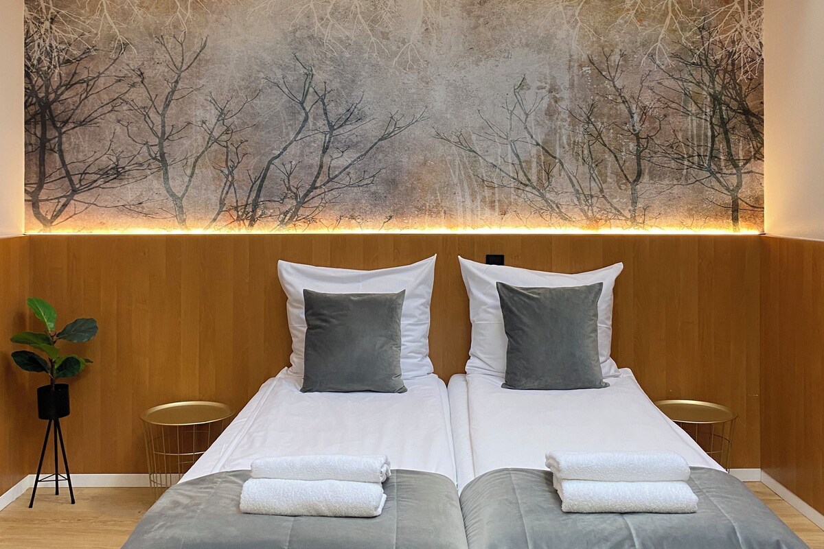 A modern hotel room with a large bed, two pillows, a bedside table, and a wall-mounted painting of bare trees.