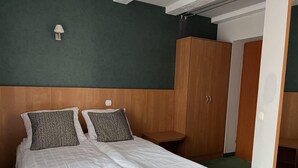 A neatly made bed with white linens and two patterned pillows, a wooden wardrobe, and a small bedside table in a room with a green wall and a white ceiling.