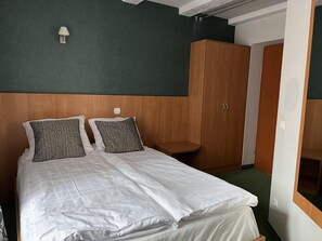Un lit bien fait, avec du linge de lit blanc et deux coussins à motifs, une armoire en bois et une petite table de chevet dans une pièce aux murs verts et au plafond blanc.