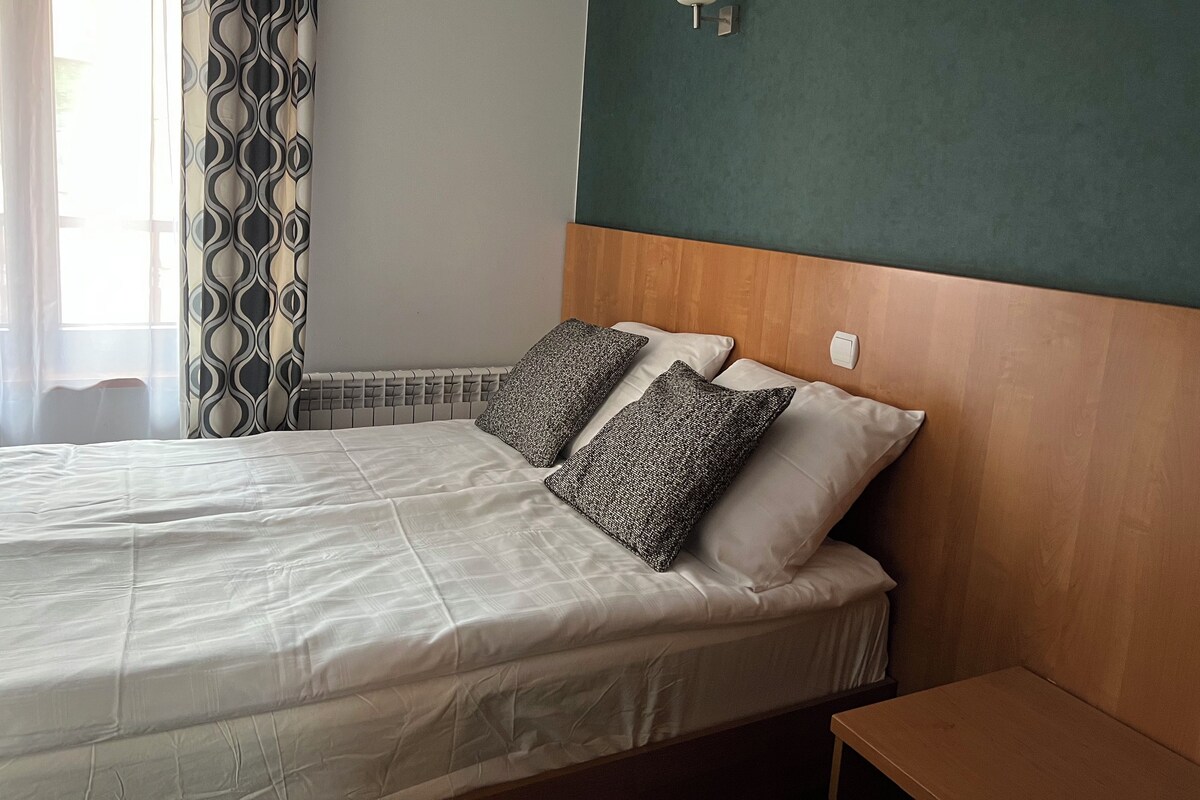 A neatly made bed with white linens and patterned pillows, a wooden headboard, and a wall-mounted light fixture.