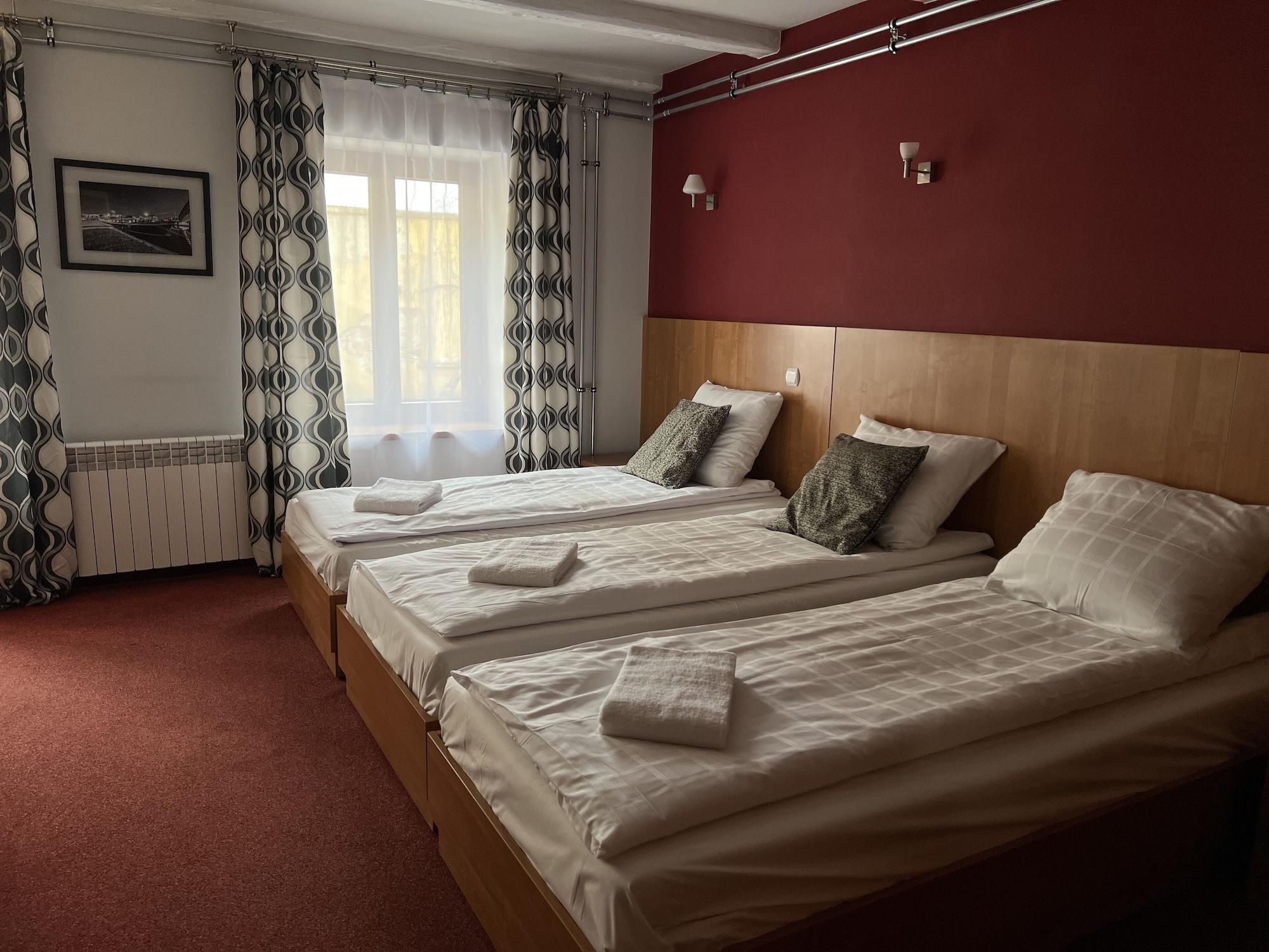 A hotel room with two beds, a red accent wall, a framed picture on the wall, and a window with patterned curtains.