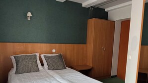 A neatly made bed with white linens and patterned pillows, a wooden wardrobe, and a small bedside table.