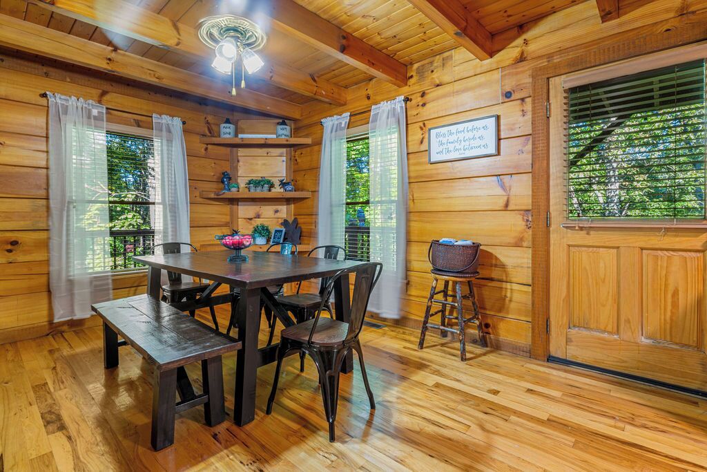 Interior de una cabaña de madera con una mesa de comedor, sillas, un banco, un ventilador de techo y una puerta con ventanal.
