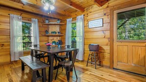 Interior de una cabaña de madera con una mesa de comedor, sillas, un banco, un ventilador de techo y una puerta con ventanal.