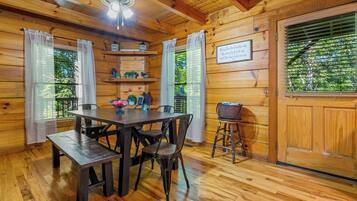 Interior de una cabaña de madera con una mesa de comedor, sillas, un banco, un ventilador de techo y una puerta con ventanal.