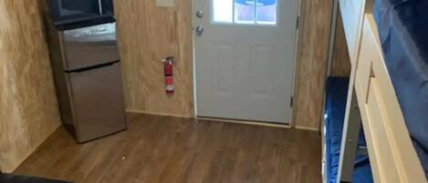 Une pièce compacte avec un plancher en bois, une kitchenette équipée d’un four à micro-ondes et d’un réfrigérateur, un lit superposé, un téléviseur fixé au mur et une porte donnant sur l’extérieur.