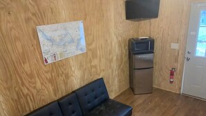 Une pièce avec un mur en bois, un canapé en cuir noir, un petit réfrigérateur, un téléviseur et une porte sur laquelle est fixé un extincteur.
