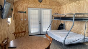 Une petite pièce comprenant un lit superposé, une table ronde en bois avec des chaises, un téléviseur et une porte avec des stores.