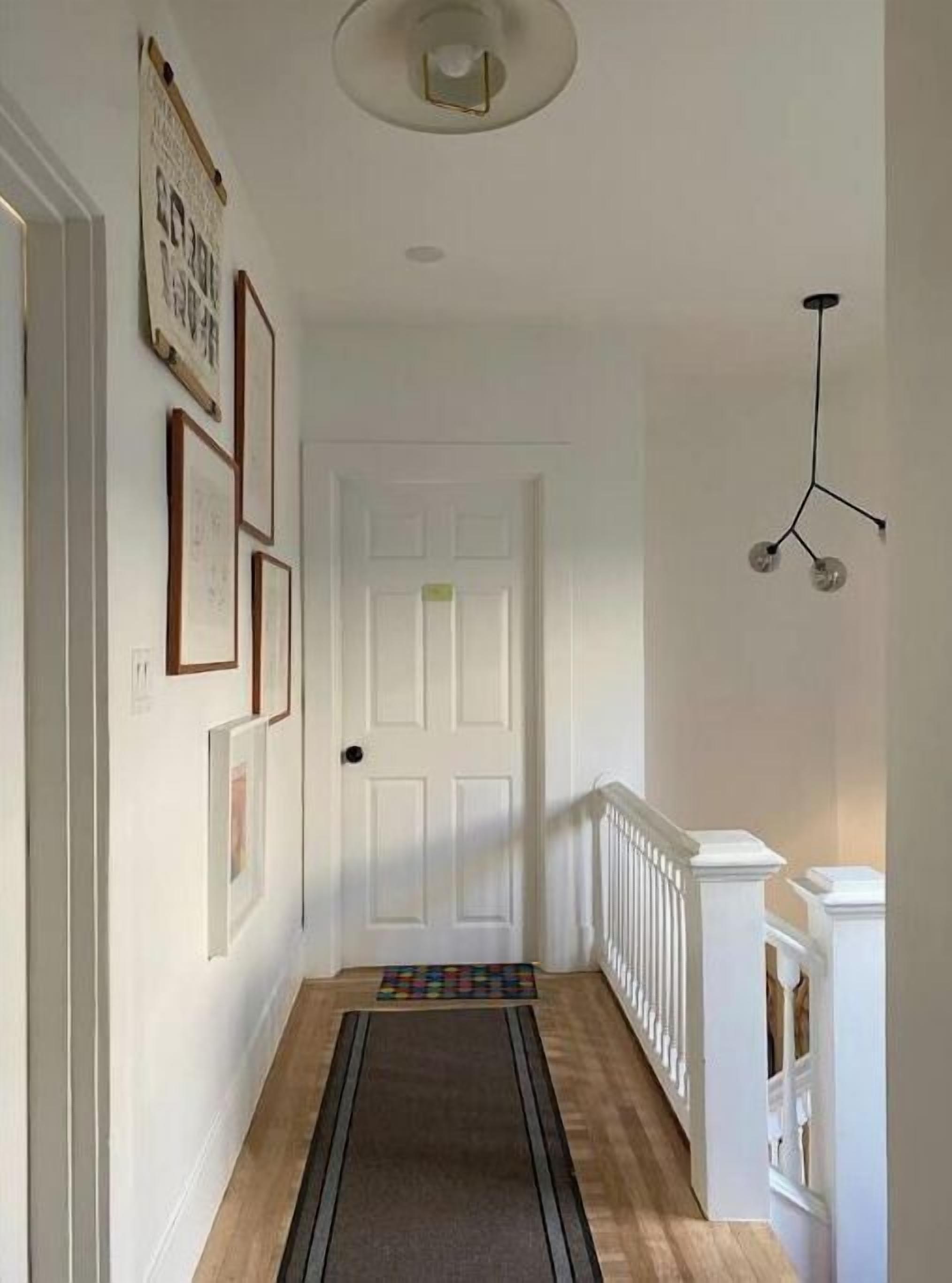 Un corridoio con pavimento in legno, pareti bianche e una porta bianca in fondo. Si notano quadri appesi al muro e un lampadario.