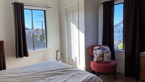 A bedroom with a bed, a chair, a window with curtains, and a view of the outside.