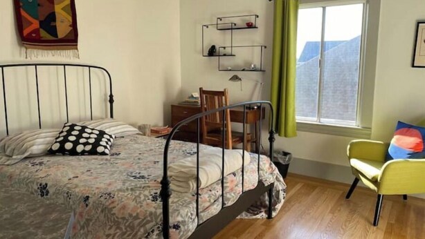 Een slaapkamer met een bed, een stoel, een bureau, een raam met gordijnen en een plank met spullen.