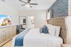 A bedroom with a bed, a ceiling fan, a TV, a dresser, and a door leading to another room.