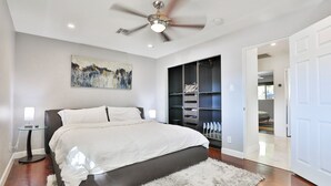 Un dormitorio con cama, ventilador de techo, un cuadro, armario y puerta que da a otra habitación.