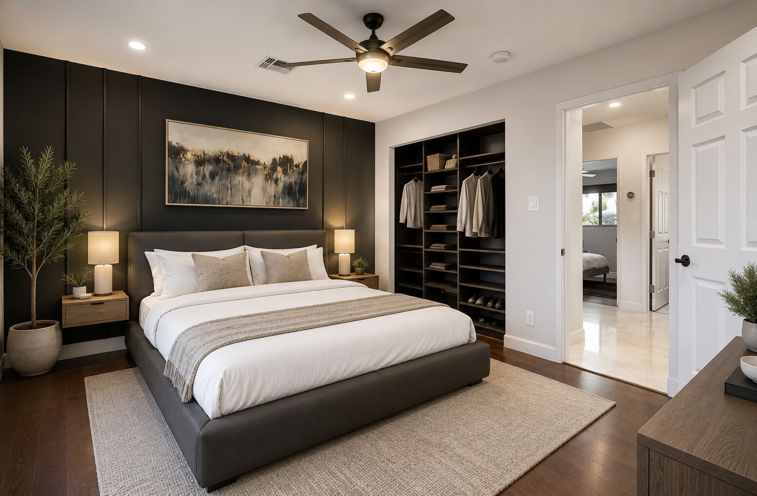 A bedroom with a large bed, a ceiling fan, a nightstand with a lamp, a wall-mounted shelf with clothes, and a closet.