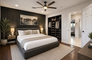 A bedroom with a large bed, a ceiling fan, a nightstand with a lamp, a wall-mounted shelf with clothes, and a closet.