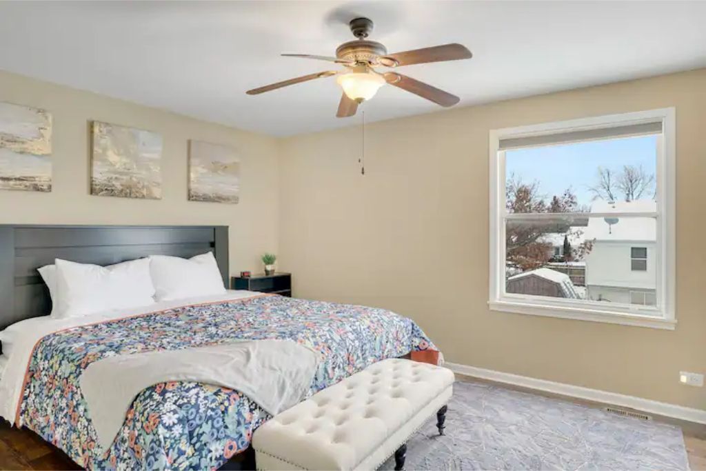 Une chambre à coucher avec un lit, un ventilateur de plafond, une fenêtre et des œuvres d’art accrochées au mur.