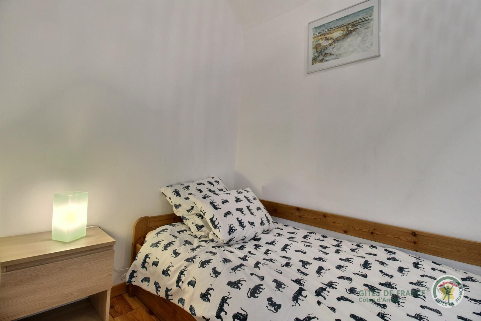 A small, neatly made bed with a patterned bedspread, a bedside table with a lamp, and a framed picture on the wall.