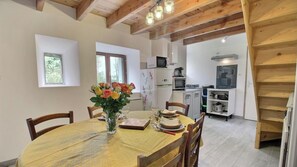 Uma sala de jantar com uma mesa redonda posta para quatro pessoas, cadeiras de madeira, um vaso com flores e uma cozinha ao fundo.
