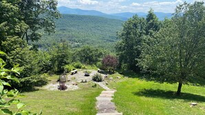 A stone pathway leading to a circular seating area with benches and a central fire feature, surrounded by lush greenery and trees. - Amazing Mountain View’s from every room! Hot Tub/3 Fire pits & Game Rm (Windham)