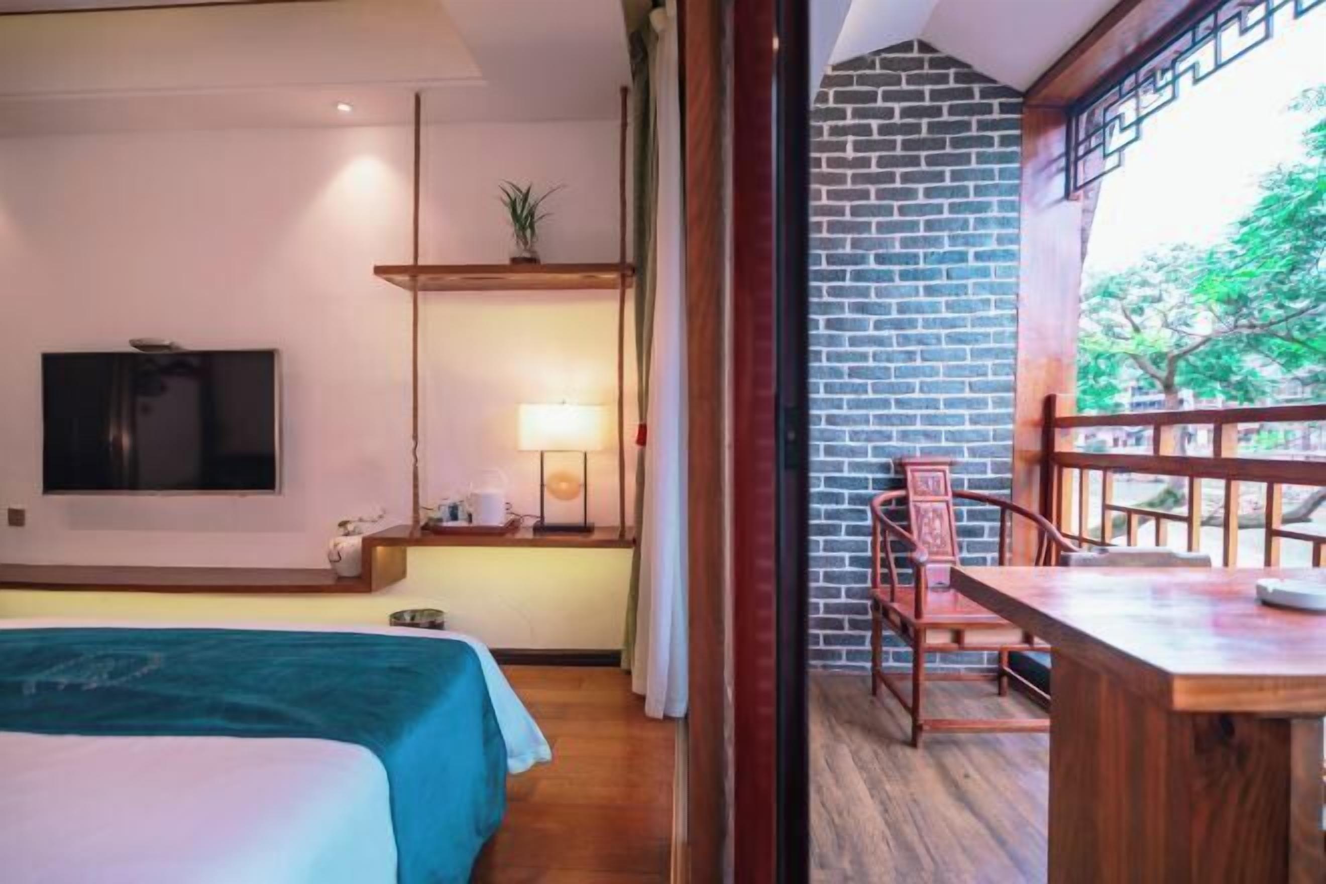A hotel room with a bed, a TV, a desk with a lamp, and a balcony with a brick wall and wooden railing.