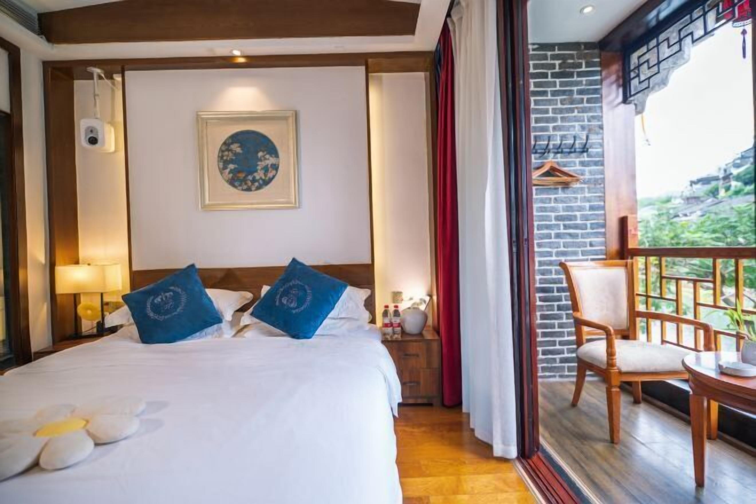 A hotel room with a bed, a nightstand, a framed picture on the wall, a balcony with a chair, and a view of greenery outside.
