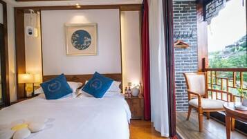 A hotel room with a bed, a nightstand, a framed picture on the wall, a balcony with a chair, and a view of greenery outside.