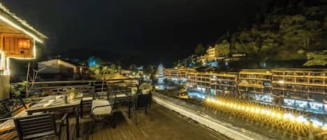 A wooden deck with tables and chairs, overlooking a lit-up river with buildings on the opposite bank.