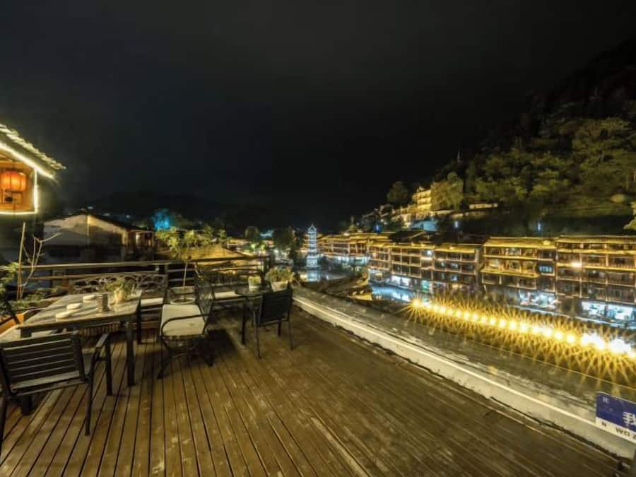 Una terraza de madera con mesas y sillas, con vistas a un río iluminado y edificios al otro lado.