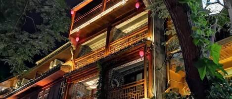 Un bâtiment traditionnel à plusieurs étages, doté de balcons en bois richement décorés et de fenêtres à croisillons, illuminé par un éclairage chaleureux.