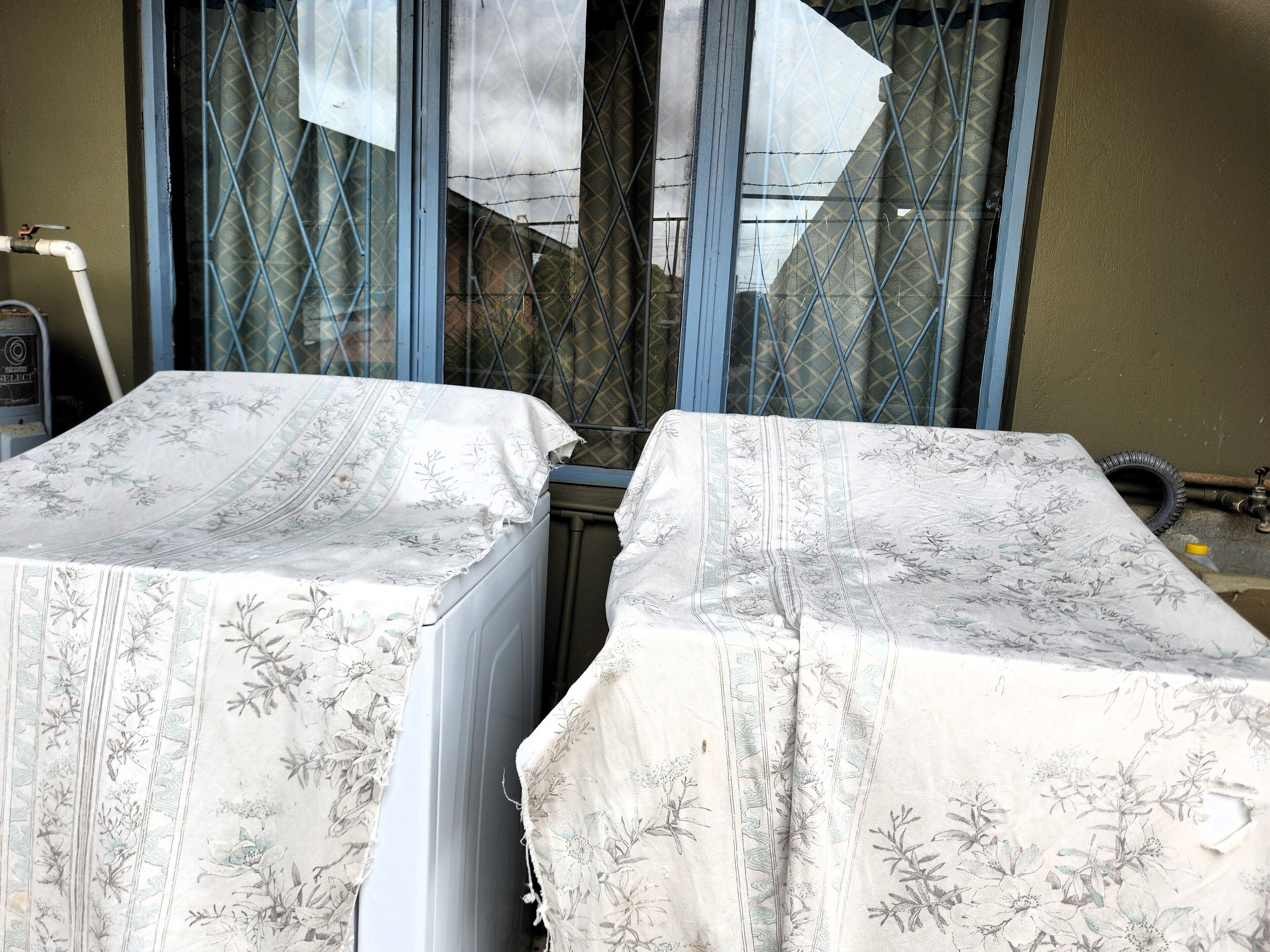 Two beds with floral-patterned covers, a window with metal bars, and a visible water pipe.