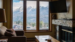 A room with a large window offering a mountain view, a flat-screen TV mounted above a fireplace, and a wooden table with papers and a remote control. - Kendall 344: Cozy Studio for 2 in the Heart of Purgatory Resort (Durango)