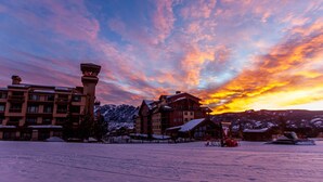 Un pueblo cubierto de nieve con edificios y un telesilla, bajo un cielo al atardecer lleno de color.