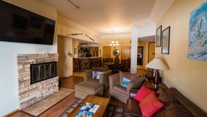 Una sala de estar con chimenea de piedra, un televisor de pantalla plana, una mesa de centro con revistas y un sofá de cuero marrón con cojines rojos. Se observa un comedor al fondo.