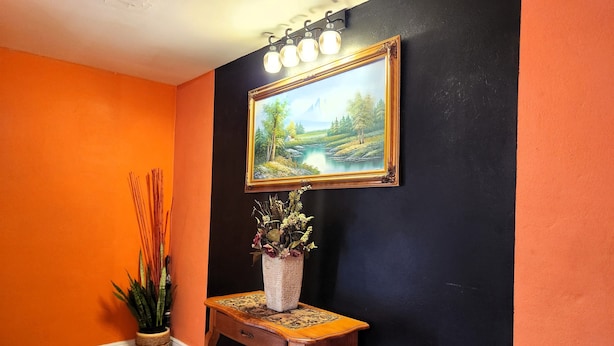 A hallway with a black wall, an orange wall, a wooden bench with a vase of flowers, and a painting above the bench.
