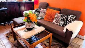 A living room with a brown sofa, patterned cushions, a wooden coffee table with a magazine, and a vase with orange flowers.