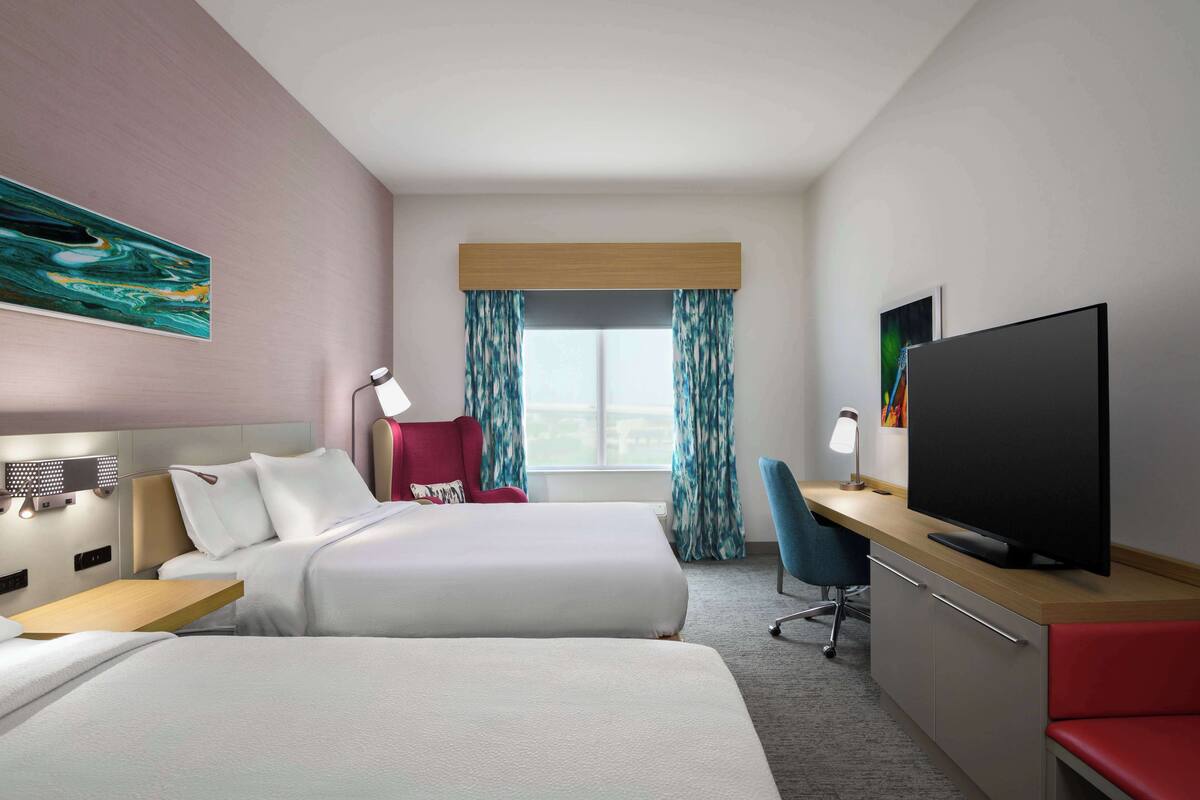 Ein Hotelzimmer mit zwei Betten, einem Schreibtisch mit Fernseher, einem Sessel, einer Schreibtischlampe und einem Fenster mit Vorhängen.