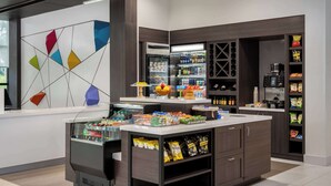 Un hall d’hôtel moderne avec un comptoir central, un réfrigérateur rempli de boissons et des étagères contenant des collations et des condiments.