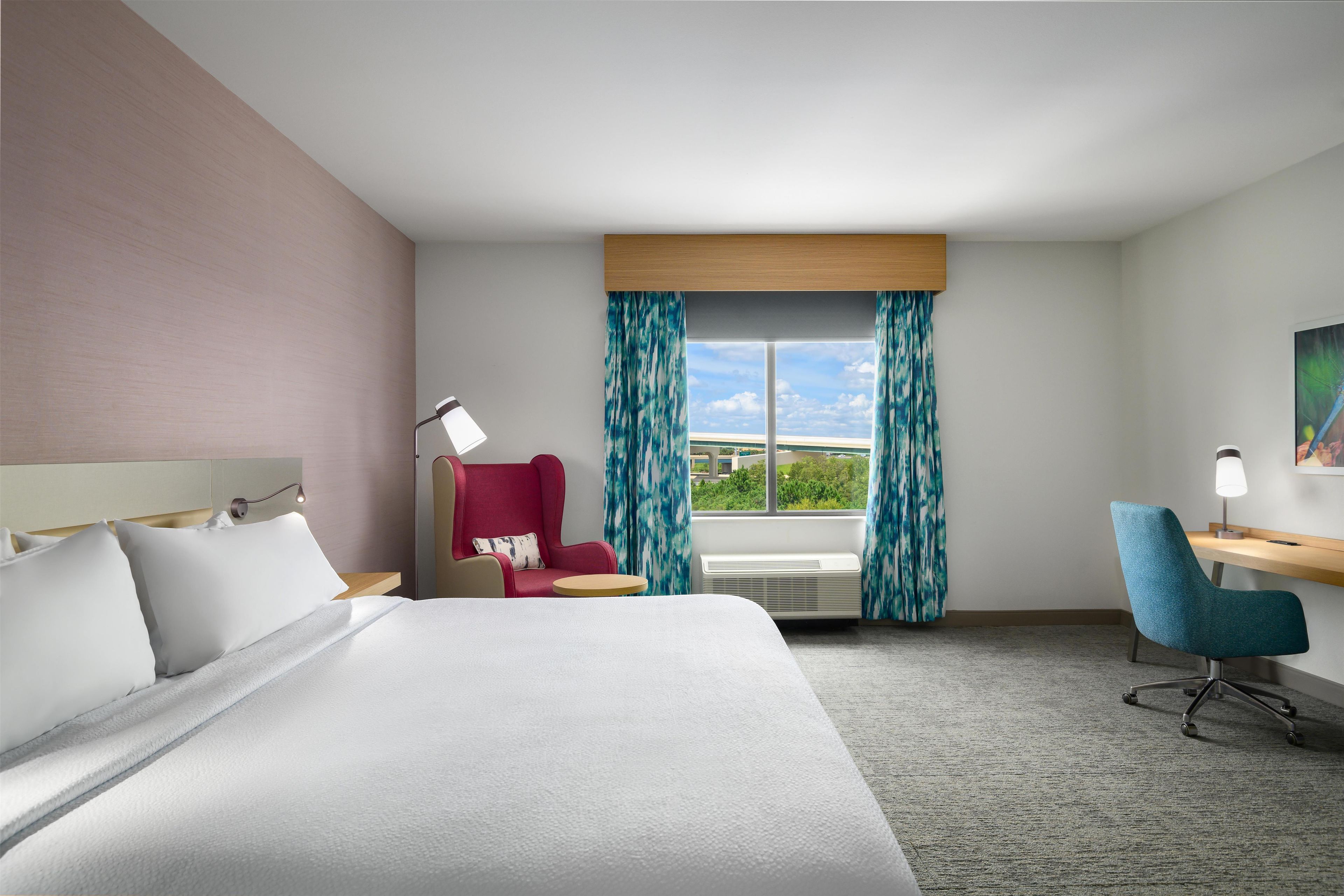 Ein Hotelzimmer mit Bett, Schreibtisch, Stuhl, einem Fenster mit Vorhängen und einem Bild an der Wand.