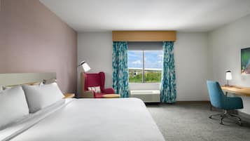 Habitación de hotel con cama, escritorio, silla, ventana con cortinas y un cuadro en la pared.