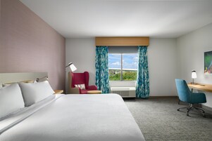 A hotel room with a bed, a desk, a chair, a window with curtains, and a painting on the wall.